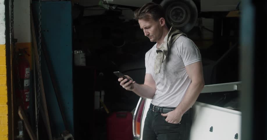 A man uses a smartphone with one hand while leaning against a vehicle in a car repair garage