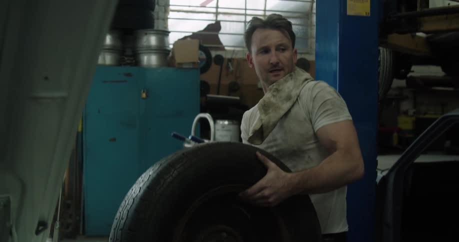 A man lifts and carries a removed car wheel in a garage