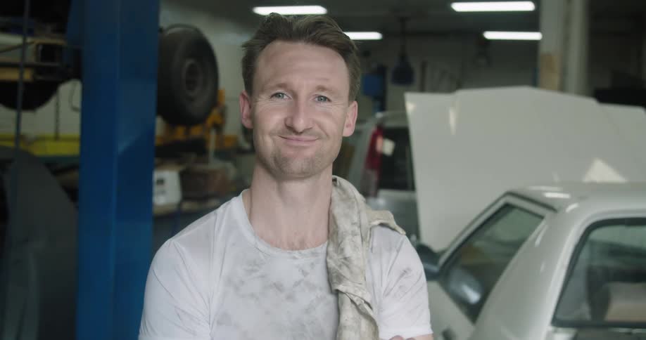 Car mechanic with a dirty white t shirt faces the camera with eye contact and smiles broadly in the garage