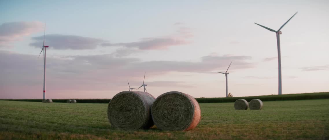 Pairs of hay bales dot the fields of a wind farm in the evening