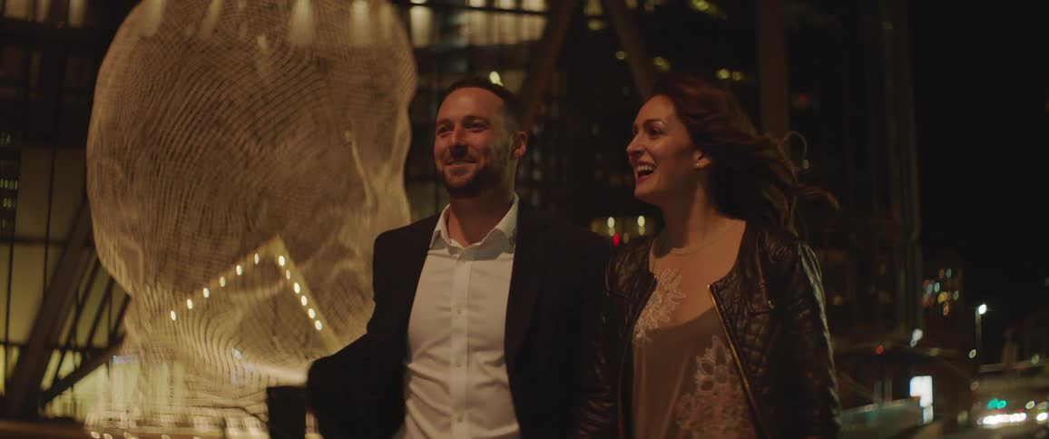 A man and woman holding hands laugh as they walk in a downtown area at night