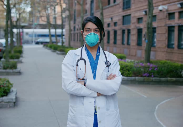Doctor with lab coat stethoscope and mask stands with arms crossed in front of building