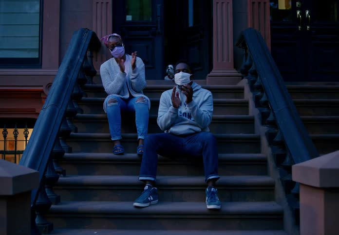 Two people wearing masks sit on steps of city home clapping