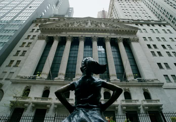 A statue of a girl with her hands on her hips stands defiantly in front of a tall building in a metropolis
