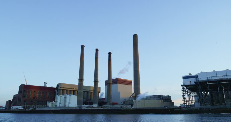 A factory with chimneys with a river in the foreground with a wind turbine and the sky in the background