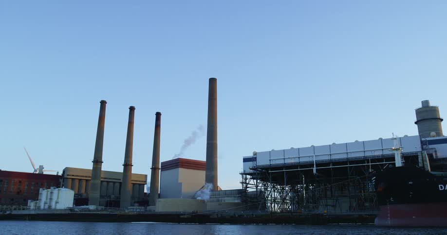 Tall smokestacks loom over a factory on the coast during the day