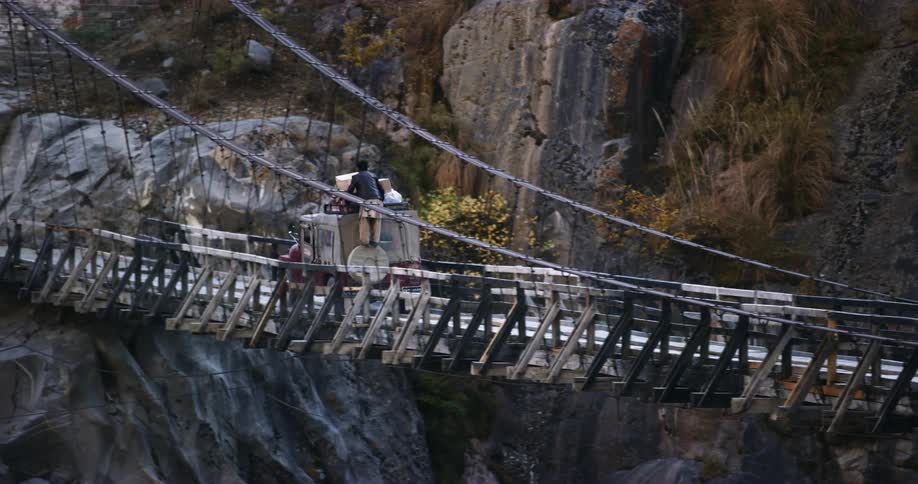 A man rides on the back of a vehicle as it travels across a wobbling wooden bridge in the mountains