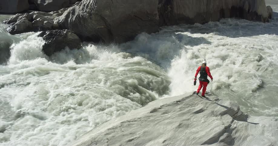 A person stands at the edge of a large boulder over rough river rapids