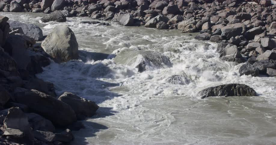 Water flowing over large rocks in a river lined with stones on a sunny day