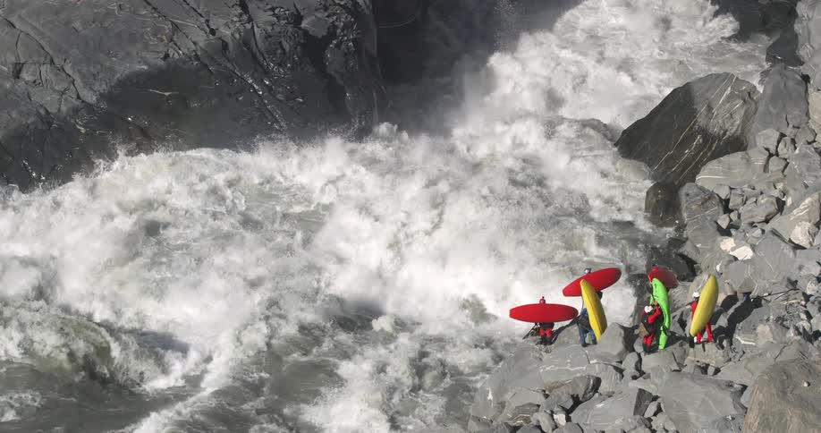 White water flows and rushes past a group of people holding colorful kayaks on the rocks