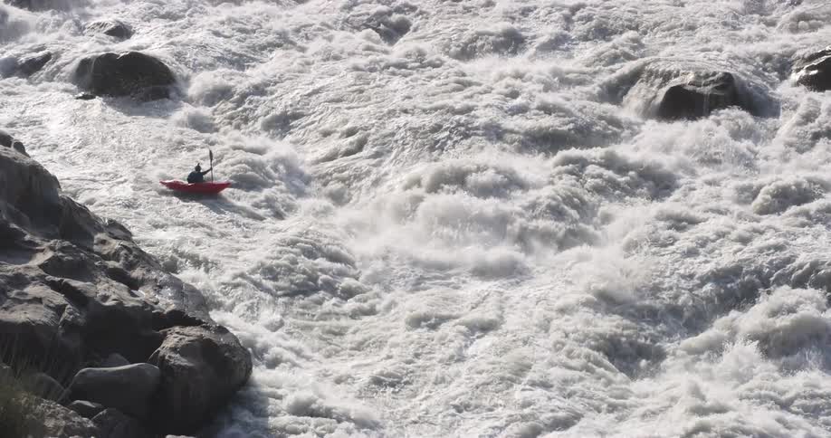 Person paddles kayak down whitewater rapids past stone strewn shore