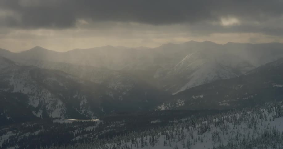 Sun rays punch through heavy clouds turning mountains and snow covered forest gloomy