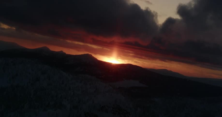 Sun sits at horizon turning sky red over mountains while snow covered forest remains dark