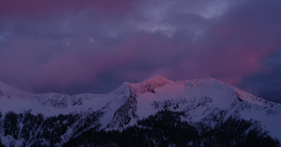 Red sunlight shines onto a snow capped mountain ridge before fading as clouds pass overhead