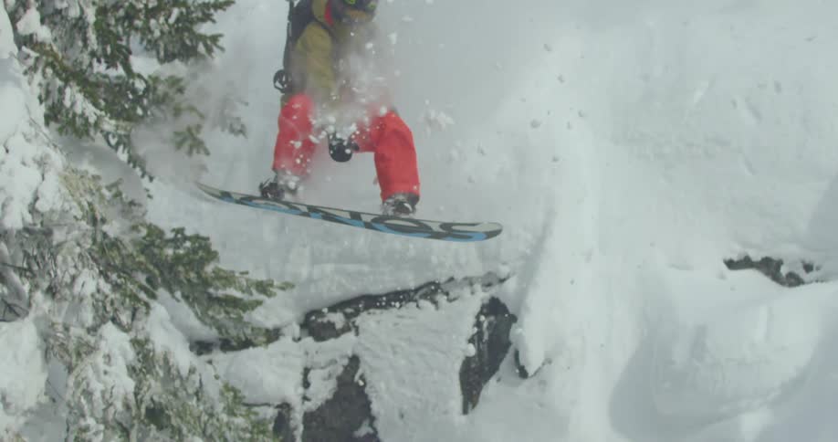 A snowboarder jumps down a steep rocky slope landing in deep fresh powder