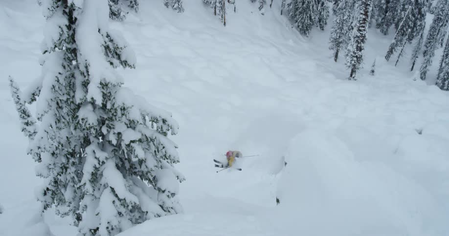 Skier wearing bright yellow clothes jumps down the snow covered cliff in the mountains near the pine tree forest