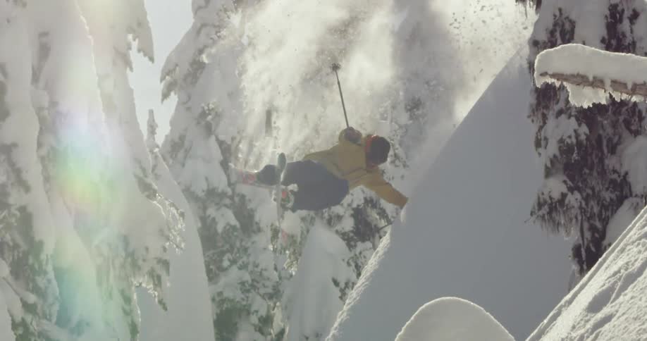 Skier wearing a yellow jacket twirls as he jumps down mountain slope past snow covered conifers as sun glares