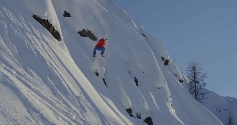 A snowboarder spins in the air as he jumps down a steep slope