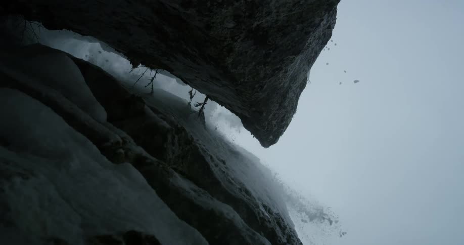 A skier rides off of a snowy rock ledge while snow sprays and falls onto the camera