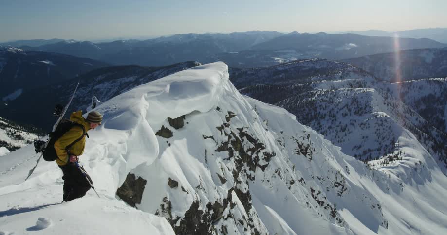 Man with snowboard on back stands on top of mountain peak