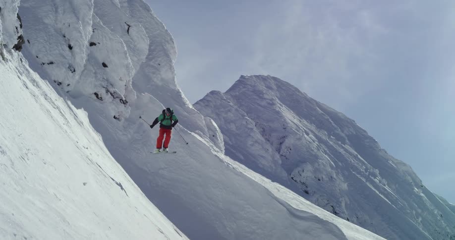 A skier jumps off a rocky mountain ledge landing on a steep snow covered slope