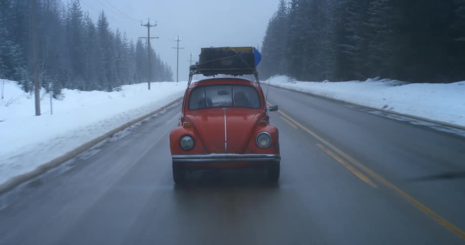 An old fashioned vehicle with luggage strapped to the top hurtles down a road through a snowy evergreen forest