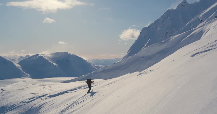 Skier walks up snow covered mountain slope using poles under lightly cloudy sky