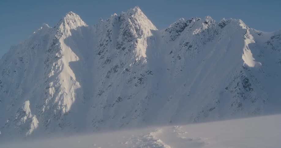 Wind blows over the snow on the plain next to the peaks of a mountain range