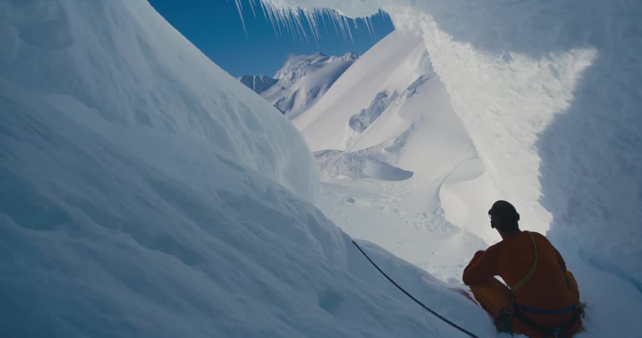Man sits in ice and snow lined crevice looking toward azure blue sky