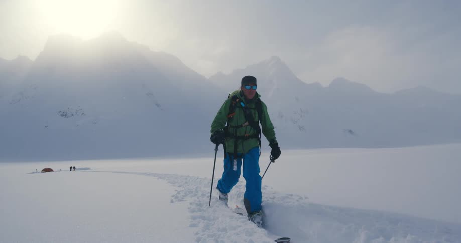 A person with a hat a backpack and sunglasses hiking on snow with skis