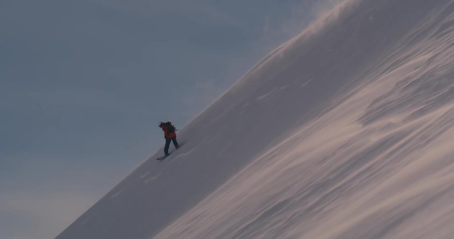 A person with a backpack snowboarding down a mountain with the sky and clouds in the background