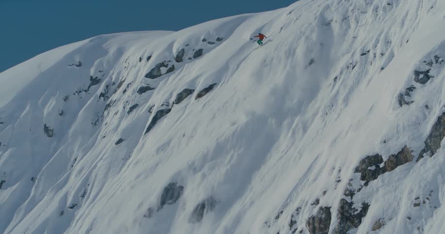A skier pauses at the top of a steep rocky ridge before skiing down and falling and tumbling