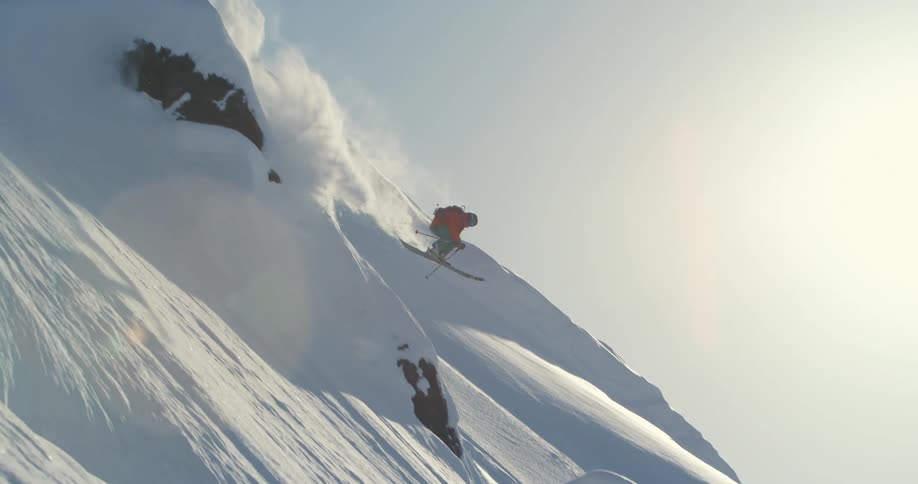 A skier navigates around stones and ice on a steep mountain slope on a sunny day