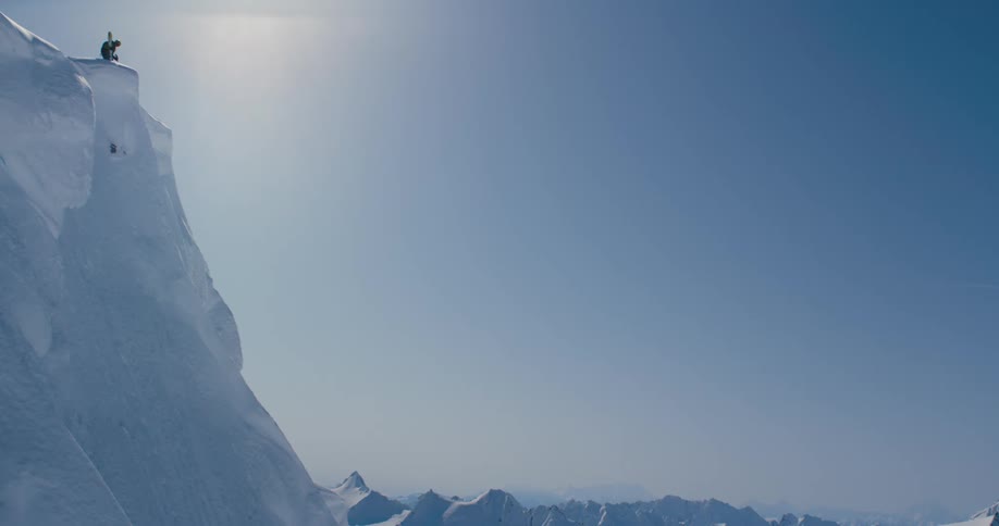 Skier stands near edge of ledge leading to steep slope on snow covered mountain on bright sunny day