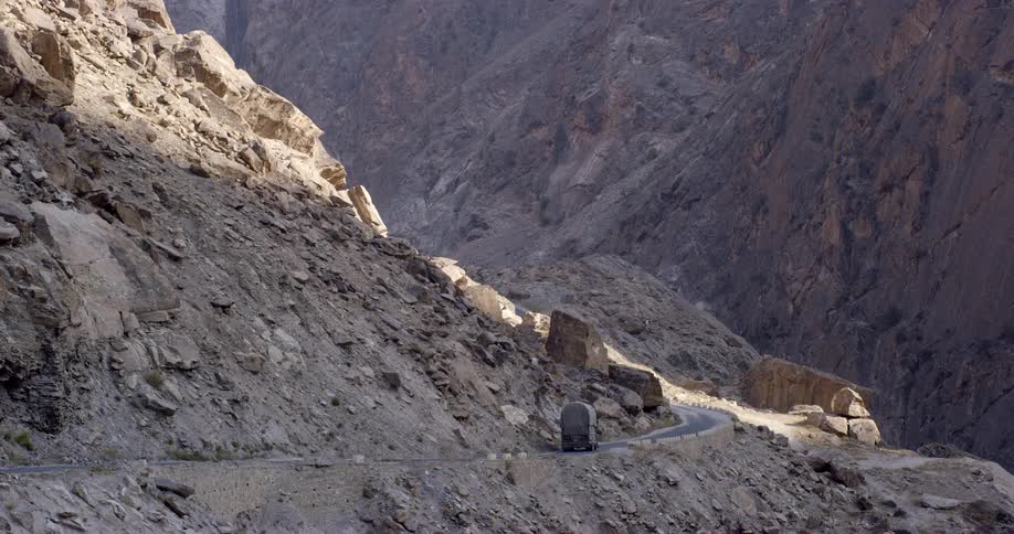 A freight truck is driving slowly down a narrow road through mountains and rocky terrain