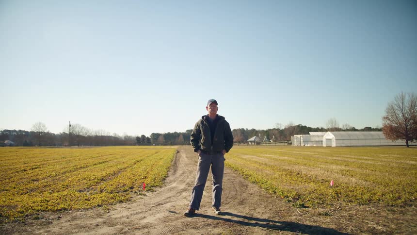A white man wearing a jacket blue jeans and a hat stands on a road leading to open farmland with greenhouses and other buildings