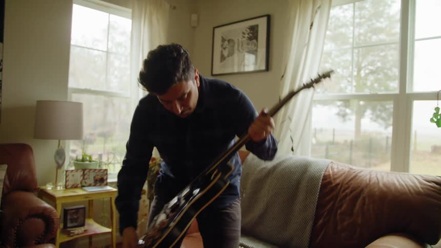 A man walks to his couch and picks up his guitar and sits down