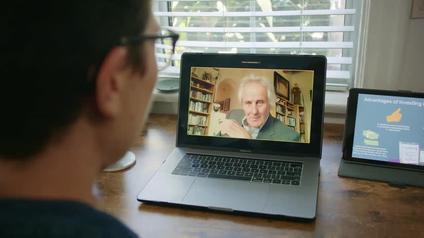 A male sits at a desk drinking from a mug and having a video call with an elderly man