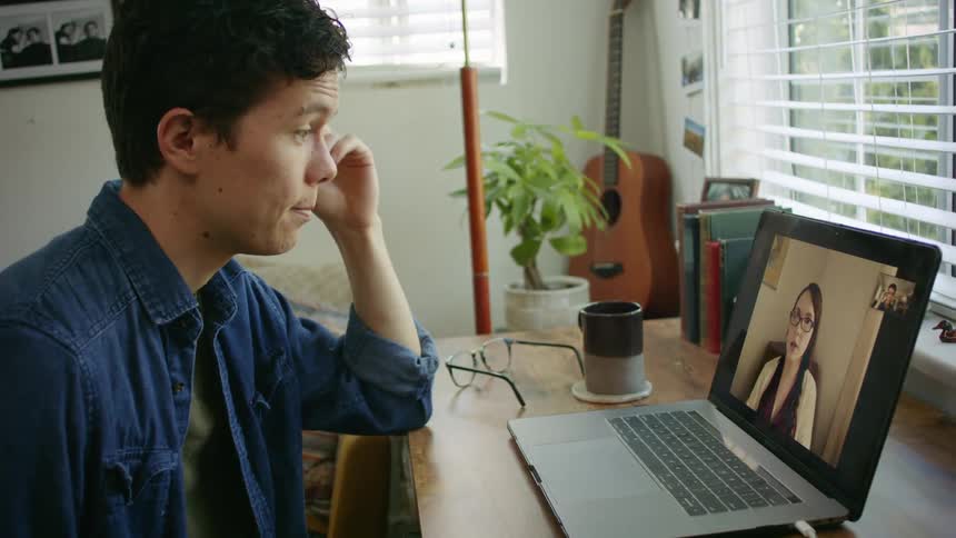 A man sits at a desk while conducting a video chat via his laptop