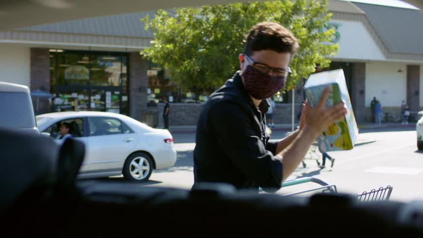 A man unloads groceries from a shopping cart into his car in a store parking lot