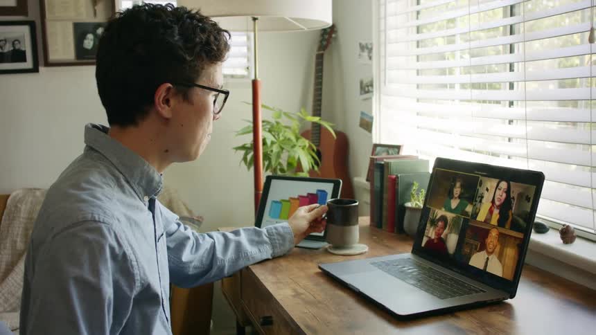 A man sits at a small desk having a video call with multiple people on a laptop
