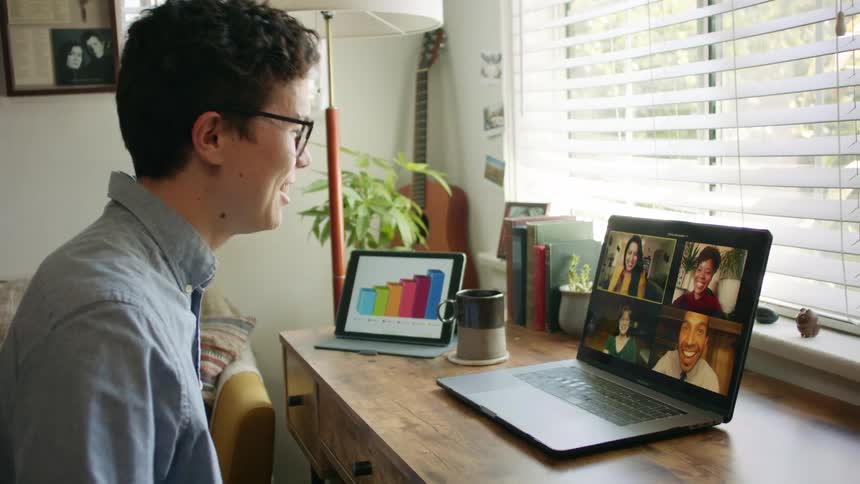 A laughing young man sits at a desk while using video chat on his laptop to speak with four other people