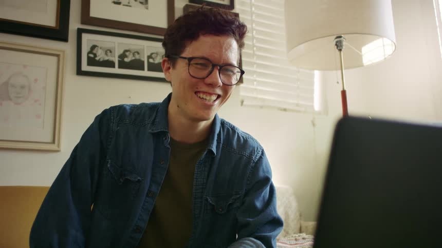A teenage boys at home waves at a laptop as he video chats