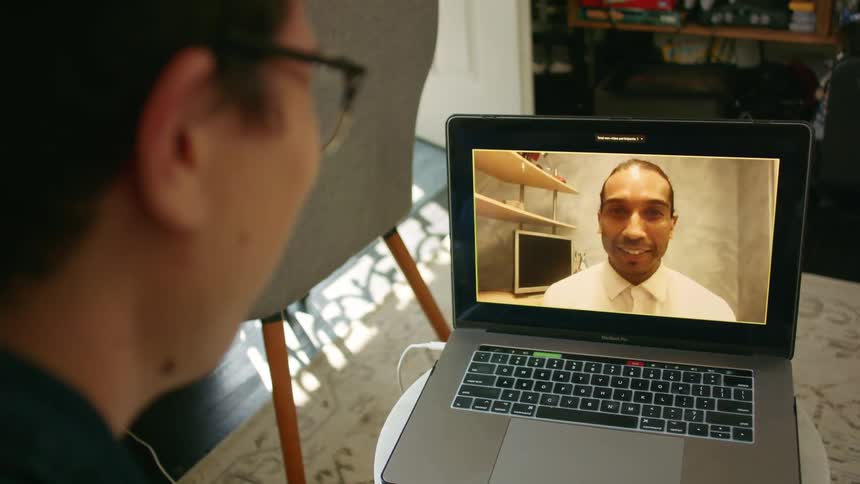 Side view of a man with glasses making a video call with a black man and waving at each other in the living room at home