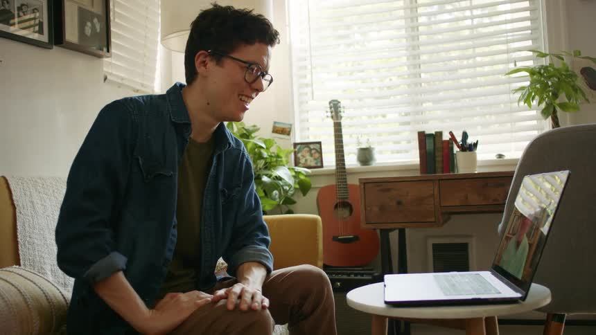 A man sitting in a couch in front of a laptop on a stool video chatting