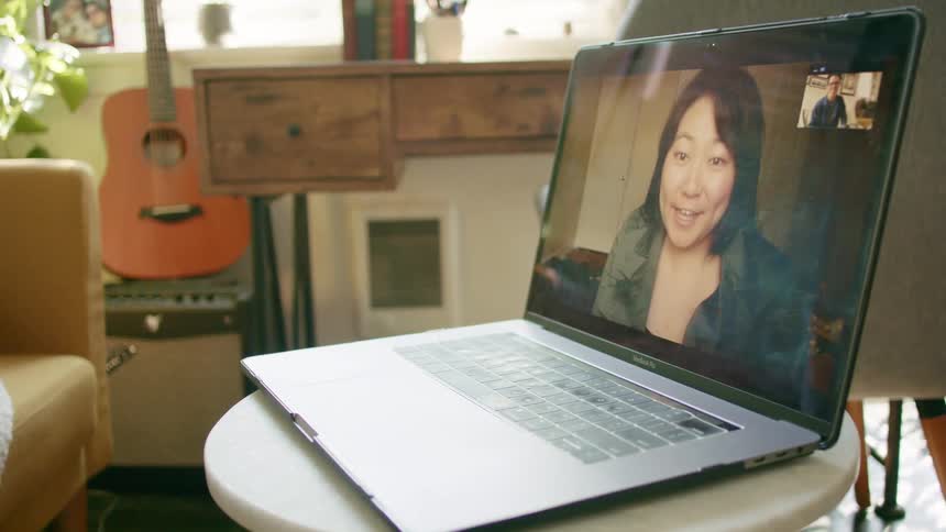 A laptop sits on a table in a house and an asian woman on the screen waves to a young man as they video chat