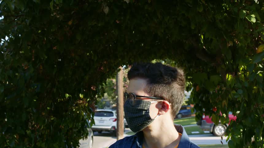 Man wearing glasses and face mask stands under arbor of vines in neighborhood