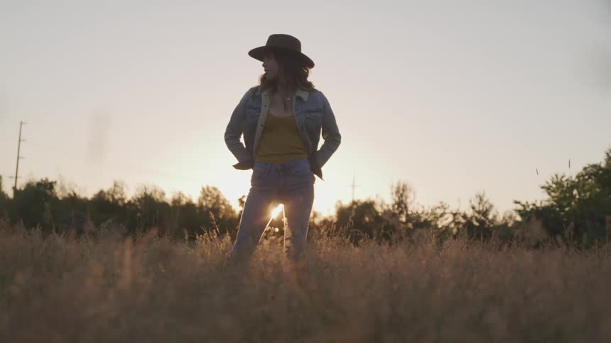A woman with a hat and jeans jacket standing in a grassy field with trees and the sky in the background