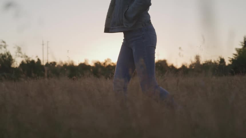 Woman with jeans and denim jacket walks through long dry grass in the field at sunset