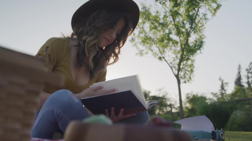 A woman with a hat sitting and reading a book with trees and the sky in the background